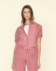 A woman wearing a matching red and white vertical-striped Xirena Channing Shirt and pants. The shirt, made of lightweight woven cotton poplin, has short sleeves and a collar, paired with a white undershirt. She stands against a plain beige background with a neutral expression.