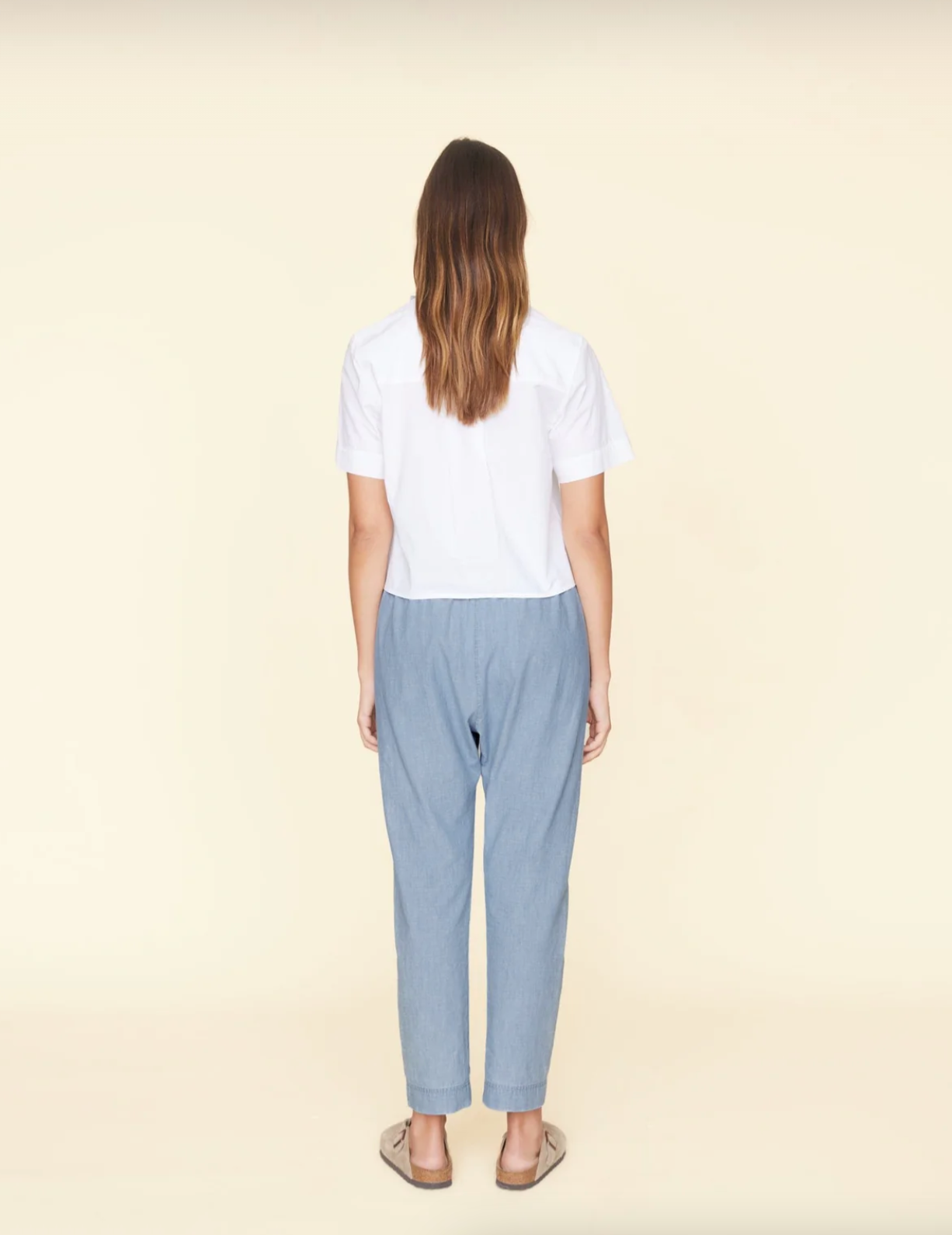 A person with long brown hair is standing with their back facing the camera against a beige background, echoing a relaxed Arizona style. They are wearing a white short-sleeved shirt, Xirena Draper Pants, and light brown slip-on shoes.