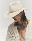 A person with shoulder-length hair is wearing a wide-brimmed, light-colored **Janessa Leone SHERMAN HAT** tilted downward, partially obscuring their face. Displaying a timeless design, the hat offers UPF 45+ protection. They have a light-colored, deep V-neck blouse with long sleeves. The background is softly lit and minimalistic.