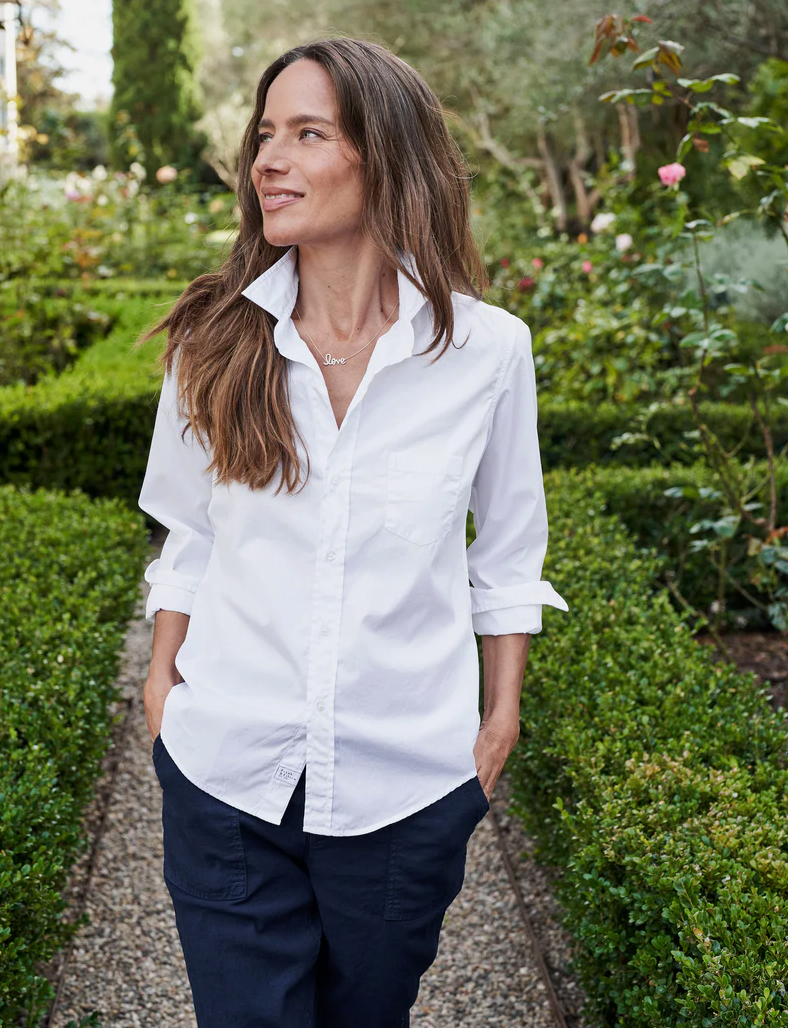 A person with long brown hair is strolling down a garden path, hands in pockets. They are wearing a white BARRY Button Up Shirt by Frank & Eileen with rolled-up sleeves and dark pants. The lush greenery and various blooming flowers give the garden an inviting, bungalow-style charm reminiscent of Arizona's serene landscapes.