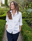 A person with long brown hair is strolling down a garden path, hands in pockets. They are wearing a white BARRY Button Up Shirt by Frank & Eileen with rolled-up sleeves and dark pants. The lush greenery and various blooming flowers give the garden an inviting, bungalow-style charm reminiscent of Arizona's serene landscapes.