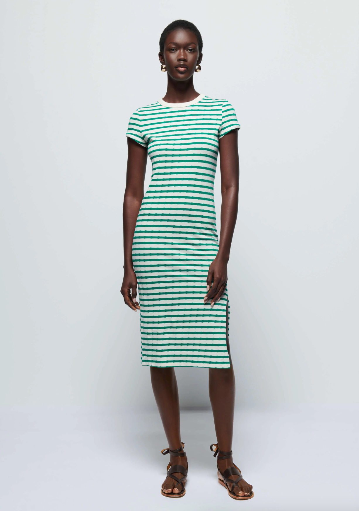 A person is standing against a plain background, wearing an Ingrid Shrunken T-Shirt Dress in Verdant Green Stripe with horizontal black stripes from Nation LTD. The short-sleeved, knee-length dress is made from 100% Pima Cotton. They have short hair, are wearing hoop earrings, and have on brown sandals with ankle straps.