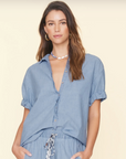 A woman with long, wavy hair is wearing a light blue Channing Shirt by Xirena made of lightweight cotton poplin, with rolled-up sleeves and matching drawstring pants. She accessorizes with gold hoop earrings and a beaded necklace. She stands against a plain beige background.