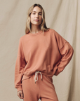 A person with long blonde hair is wearing The Teammate Sweatshirt by The Great Inc., along with matching peach-colored sweatpants, featuring vintage details, posing against a neutral fabric background. They have a relaxed expression and their left hand rests on their hip.