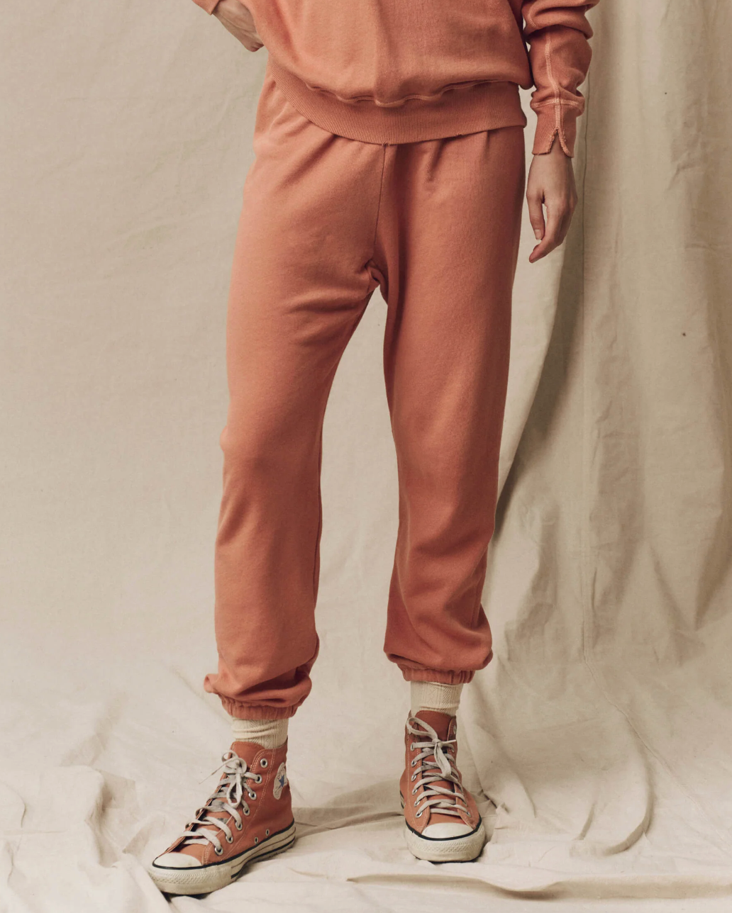 A person is wearing matching peach-colored sweatpants and a sweatshirt, all made of 100% cotton. The Stadium Sweatpant by The Great Inc. are cuffed at the ankles, revealing peach socks, and they are also wearing peach-colored high-top sneakers with white laces and soles. The background is a neutral beige fabric.