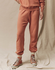 A person is wearing matching peach-colored sweatpants and a sweatshirt, all made of 100% cotton. The Stadium Sweatpant by The Great Inc. are cuffed at the ankles, revealing peach socks, and they are also wearing peach-colored high-top sneakers with white laces and soles. The background is a neutral beige fabric.