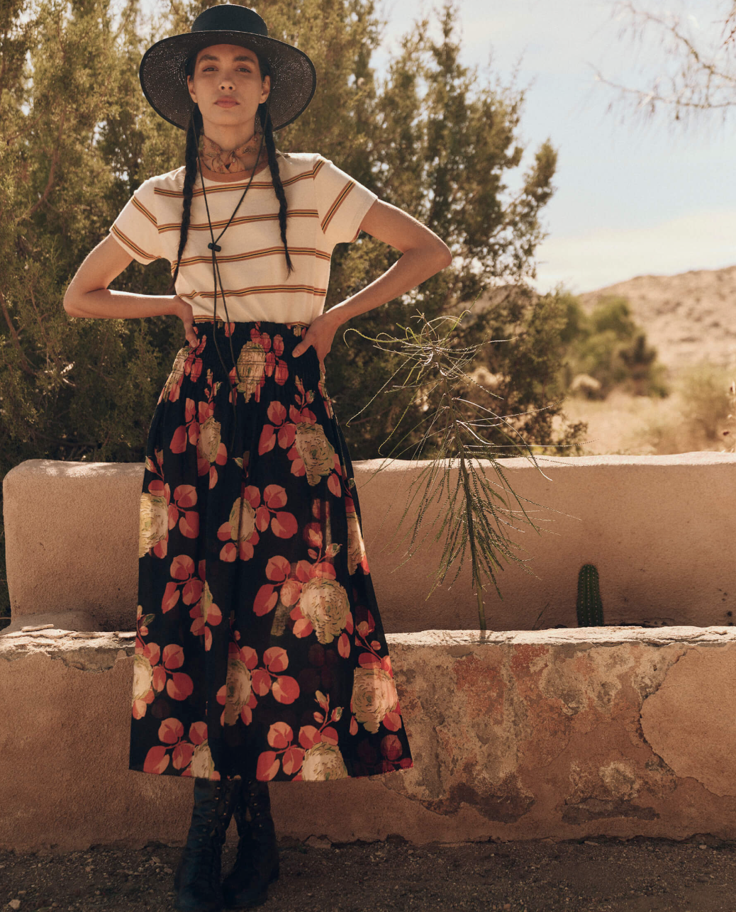 A woman with long braided hair stands outdoors, wearing a wide-brimmed black hat, The Little Tee by The Great Inc. as a layering piece, and a long floral skirt. She is positioned in front of a low wall with desert plants and bushes in the background.