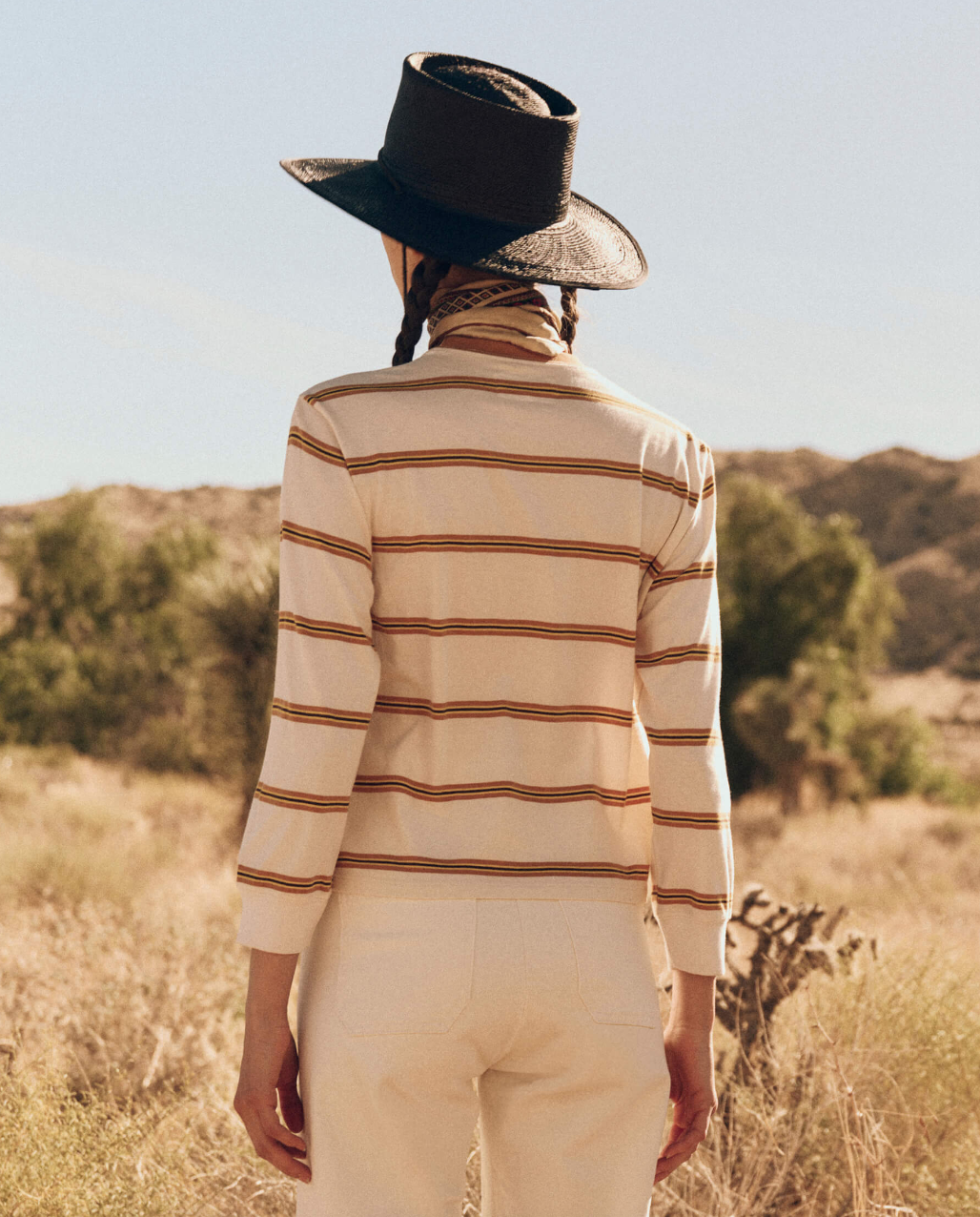 A person with braided hair stands outdoors, facing away from the camera. They wear a wide-brimmed black hat, a long-sleeve The Campus Crew shirt by The Great Inc., and light-colored pants. The landscape features dry grass and distant trees under a clear sky.