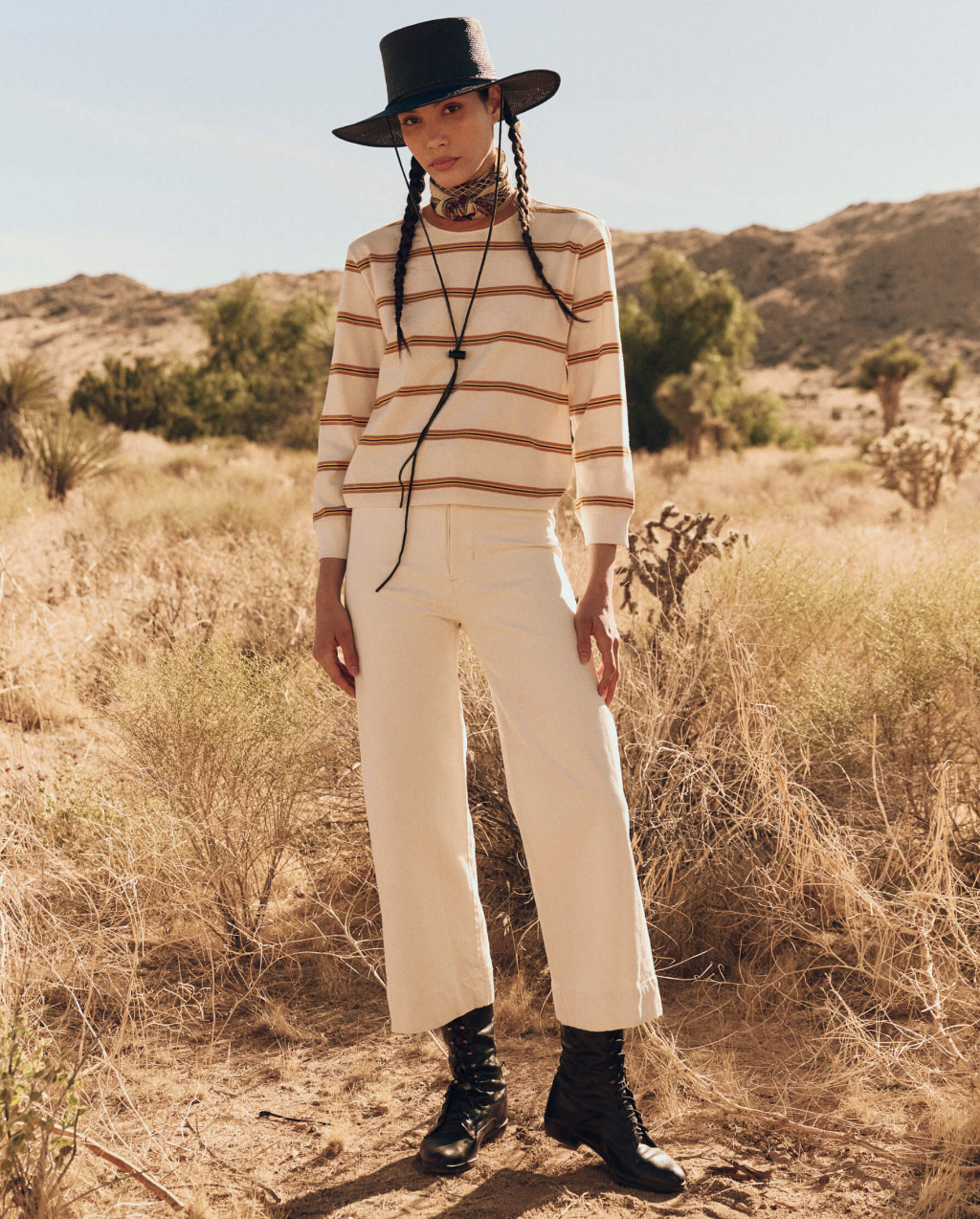 A person stands in a desert landscape wearing a wide-brimmed black hat, The Campus Crew beige and white long-sleeved shirt by The Great Inc., white pants, and black lace-up boots. They have long braided hair and look directly at the camera with a neutral expression. Sparse bushes and hills are in the background.