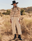 A person stands in a desert landscape wearing a wide-brimmed black hat, The Campus Crew beige and white long-sleeved shirt by The Great Inc., white pants, and black lace-up boots. They have long braided hair and look directly at the camera with a neutral expression. Sparse bushes and hills are in the background.