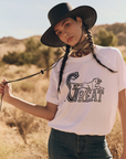 A person stands outdoors in a dry, grassy area wearing a wide-brimmed black hat, The Short Sleeve Sweatshirt from The Great Inc. with a leaping tiger graphic, a colorful neckerchief, and blue jeans. The person has two long braids and holds the hat's chin strap.