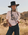 A person with long braided hair wearing a wide-brimmed black hat, a bandana around their neck, "The Short Sleeve Sweatshirt" by The Great Inc. and blue jeans stands in a dry, desert-like landscape.