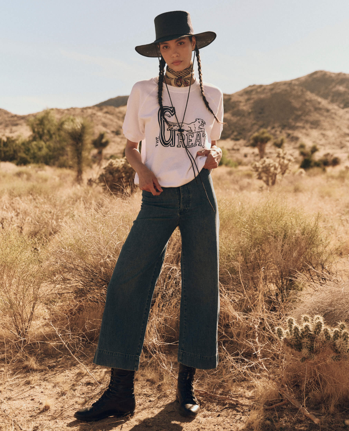 A person stands in a desert landscape wearing a wide-brimmed hat, The Short Sleeve Sweatshirt by The Great Inc., high-waisted jeans, and black boots. They have long hair styled in two braids and accessorize with a neckerchief and a belt. Bushes and mountains are visible in the background.