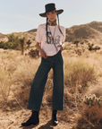 A person stands in a desert landscape wearing a wide-brimmed hat, The Short Sleeve Sweatshirt by The Great Inc., high-waisted jeans, and black boots. They have long hair styled in two braids and accessorize with a neckerchief and a belt. Bushes and mountains are visible in the background.