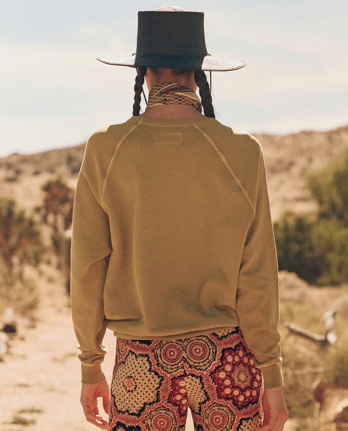 A person stands in a desert landscape with their back to the camera. They have two braids and wear a wide-brimmed hat, a vintage sweatshirt, an intricately patterned scarf around their neck, and colorful, patterned pants. The College Sweatshirt by The Great Inc. stands out against the sandy terrain and sparse vegetation.