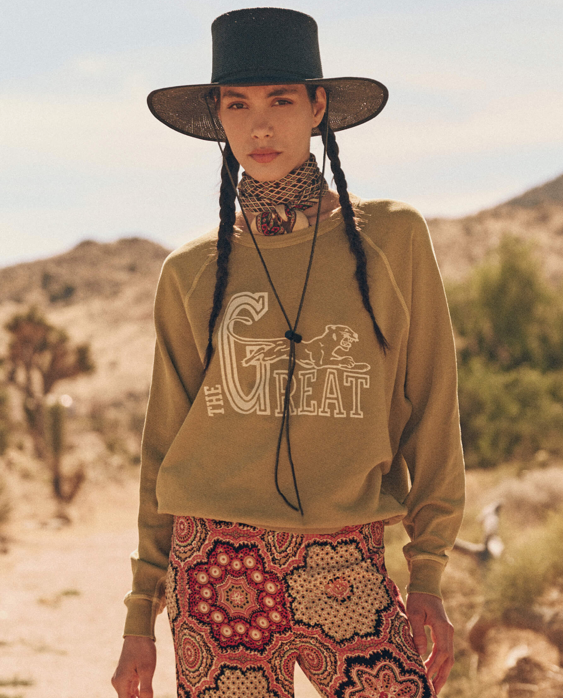 A woman stands outdoors in a desert landscape wearing a wide-brimmed black hat, a tan scarf, The College Sweatshirt by The Great Inc. with "THE GREAT" and a horse graphic, and patterned pants, capturing an effortlessly chic style reminiscent of handcrafted Los Angeles fashion.