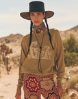 A woman stands outdoors in a desert landscape wearing a wide-brimmed black hat, a tan scarf, The College Sweatshirt by The Great Inc. with "THE GREAT" and a horse graphic, and patterned pants, capturing an effortlessly chic style reminiscent of handcrafted Los Angeles fashion.