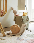 A cozy nursery room with a large, soft Cushion Lazy Snail by Lorena Canals on a patterned rug. In the background, there's a woven basket filled with plush toys, situated next to a light-colored wooden crib. Sunlight streams through a partially open window, illuminating the serene Fantasy Garden-like scene.