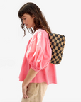 A person with long hair is wearing a pink blouse and beige pants, carrying the Petite Moyen S24 bag by Clare Vivier, which boasts handwoven leather details and crossbody straps against a plain white backdrop.