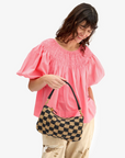 A person wearing a pink top with puffed sleeves and light-colored pants holds the Clare Vivier Petite Moyen S24, a mini messenger bag with handwoven leather. They have long, wavy hair and are smiling while looking down.