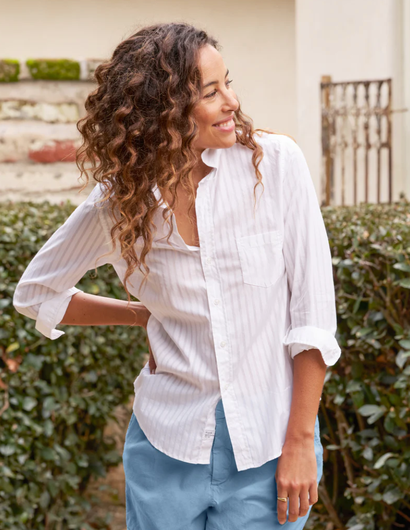 A woman with curly hair smiles while looking to the side. She is wearing a tailored fit, white, long-sleeved BARRY Button Up Shirt by Frank & Eileen and blue pants. She stands in front of a garden with trimmed hedges and a partial view of a building with a stone wall and iron gate in the background.