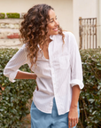 A woman with curly hair smiles while looking to the side. She is wearing a tailored fit, white, long-sleeved BARRY Button Up Shirt by Frank & Eileen and blue pants. She stands in front of a garden with trimmed hedges and a partial view of a building with a stone wall and iron gate in the background.