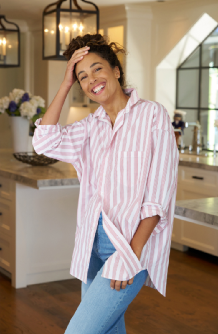 Shirley, with her curly hair, is smiling and standing in a modern kitchen. She’s wearing a pink and white striped SHIRLEY Shirt by Frank &amp; Eileen and blue jeans, with one hand on her head and the other in her pocket. The kitchen features white cabinets, pendant lights, and a vase of flowers.