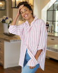 Shirley, with her curly hair, is smiling and standing in a modern kitchen. She’s wearing a pink and white striped SHIRLEY Shirt by Frank & Eileen and blue jeans, with one hand on her head and the other in her pocket. The kitchen features white cabinets, pendant lights, and a vase of flowers.