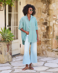 A person stands outside, wearing a green and white striped SHIRLEY Shirt from Frank & Eileen, light blue flared jeans, and sandals. They have dark curly hair and are wearing sunglasses, smiling slightly and looking to the side. The background features a stone wall and a potted succulent.