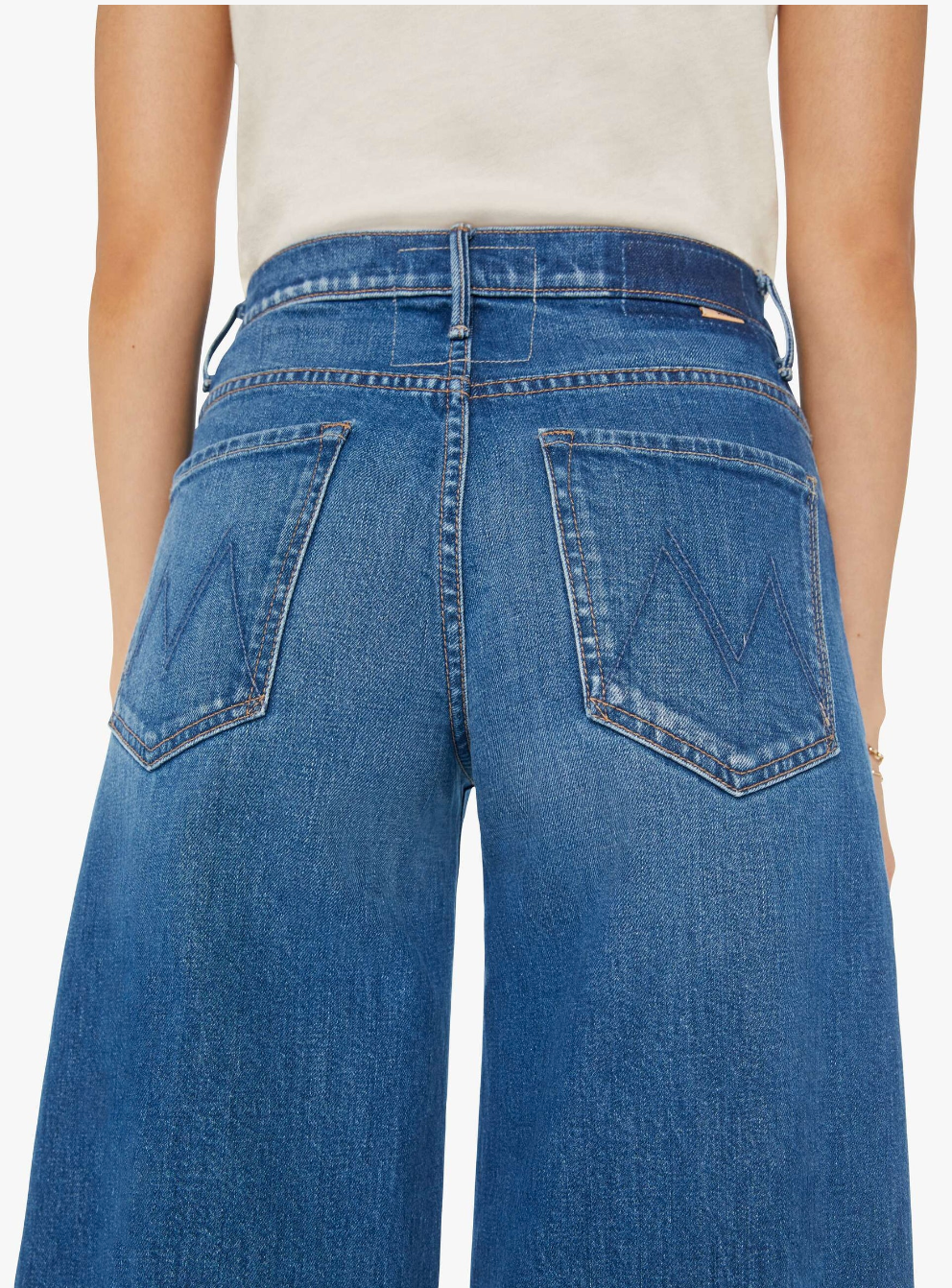 A person wearing blue denim The Swisher Sneak Fray jeans by Mother with visible back pocket stitching in an &quot;M&quot; design is shown from the waist down. They are also wearing a beige top. The high-rise jeans have a relaxed fit, are made of semi-rigid SUPERIOR denim, and feature prominent stitching details.