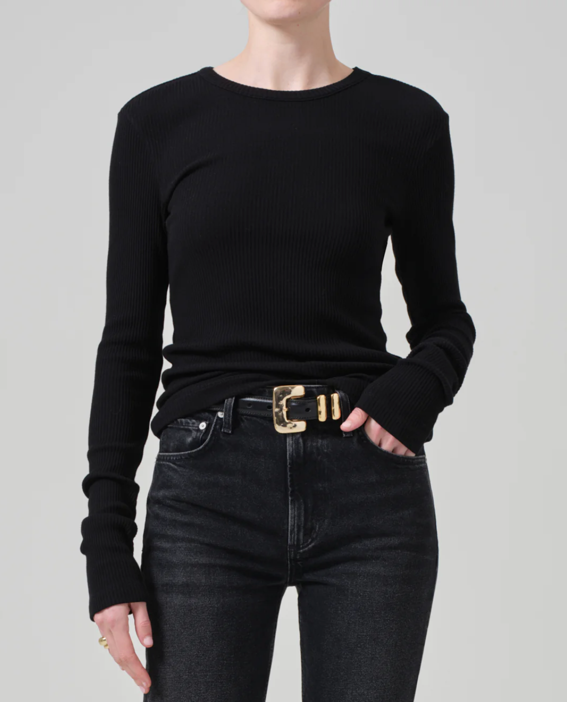 A person with short hair is wearing a fitted Bina Crewneck black long-sleeve ribbed shirt made from organic cotton by Citizens Of Humanity/AGOLDE, paired with high-waisted black jeans. They have a gold-buckled black belt and a gold ring on their right hand. The background is plain light grey.