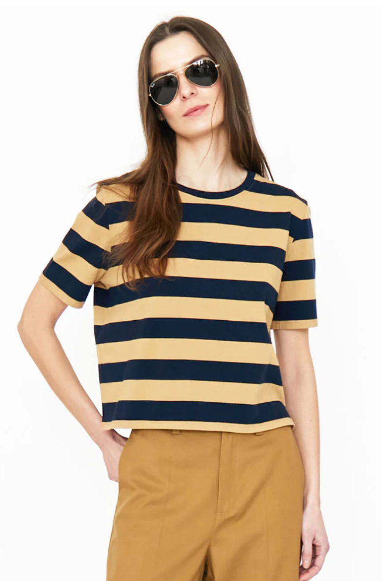 A person with long hair wearing aviator sunglasses, a short-sleeved beige and black striped modern tee called The Brentwood by Kule, and mustard-colored pants stands against a plain white background. One hand is in the pocket, and the other arm is relaxed by their side.