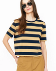 A person with long hair wearing aviator sunglasses, a short-sleeved beige and black striped modern tee called The Brentwood by Kule, and mustard-colored pants stands against a plain white background. One hand is in the pocket, and the other arm is relaxed by their side.