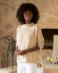 A person with curly hair stands indoors, wearing a luxurious Monterey Sweater by Frank & Eileen and white pants. They have a contemplative expression and are adorned with hoop earrings and bracelets. In the background, there is a stone wall, a decorative sculpture, a table, and various items.