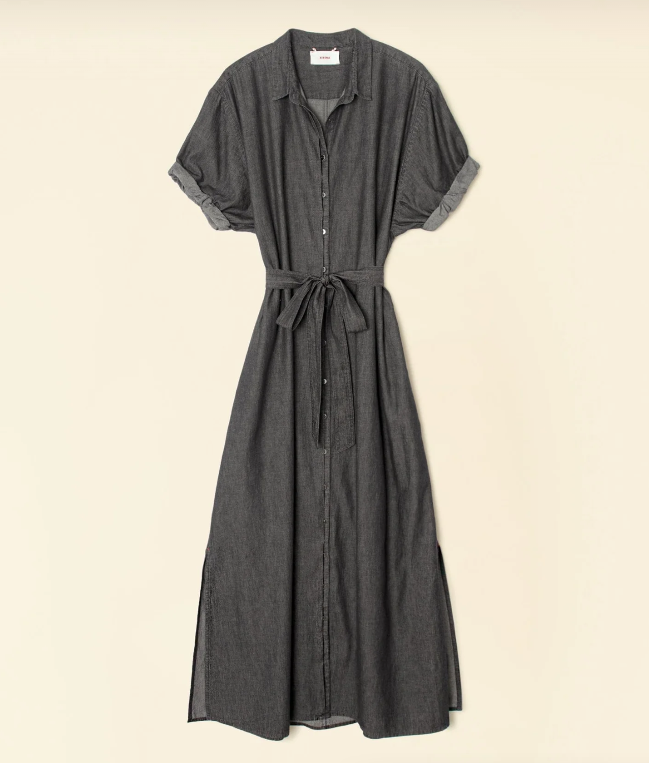 A long Linnet Dress by Xirena, crafted from dark grey denim, featuring short sleeves, a button-down front, a fabric belt at the waist, and side slits at the hem. The tailored fit dress is hanging against a plain beige background.