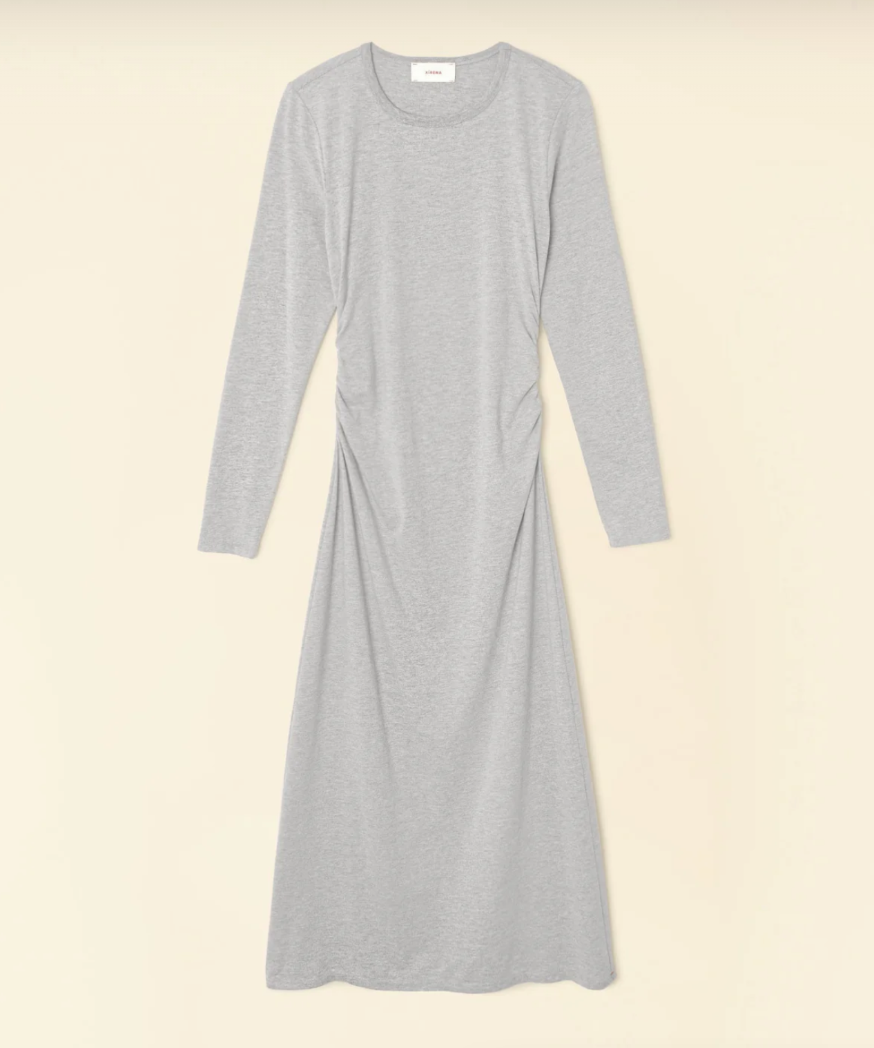 The Wiley Dress by Xirena is a light gray, long-sleeve, midi-length dress made from mid-weight cotton jersey. It has a simple round neckline and features subtle side ruching at the waist for a slightly gathered effect. The dress is laid flat on a beige background.