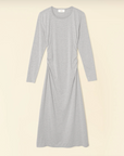 The Wiley Dress by Xirena is a light gray, long-sleeve, midi-length dress made from mid-weight cotton jersey. It has a simple round neckline and features subtle side ruching at the waist for a slightly gathered effect. The dress is laid flat on a beige background.