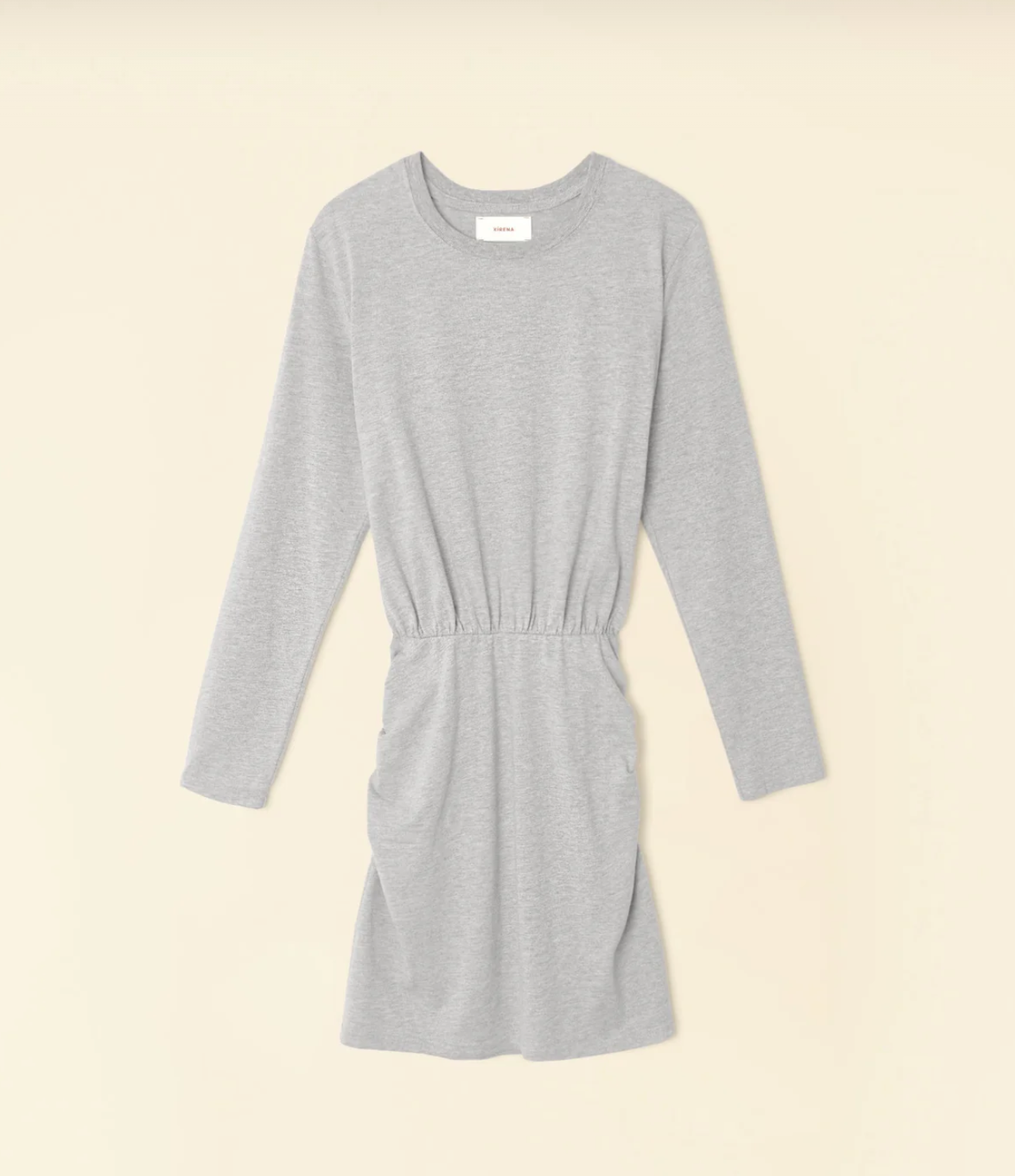 Introducing the Lula Dress by Xirena - a light gray, long sleeve mini dress crafted from soft, stretchy fabric. It boasts a round neckline and an elastic ruched waist that creates a subtle gathered effect. With its simple, casual design, this dress falls above the knee and is showcased beautifully against a beige background.