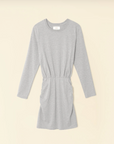 Introducing the Lula Dress by Xirena - a light gray, long sleeve mini dress crafted from soft, stretchy fabric. It boasts a round neckline and an elastic ruched waist that creates a subtle gathered effect. With its simple, casual design, this dress falls above the knee and is showcased beautifully against a beige background.