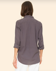 A person with long brown hair is shown from behind wearing a menswear-inspired Beau Shirt by Xirena, featuring long sleeves and a button-down design in gray cotton poplin with rolled-up sleeves. They pair the shirt with white pants, standing against a neutral beige background.