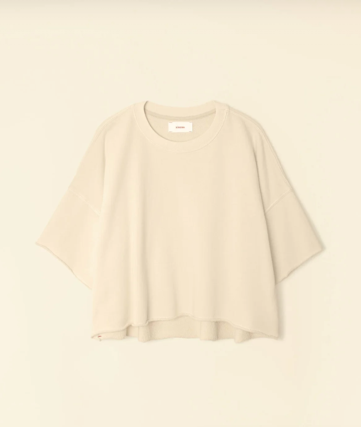 A light beige, oversized Romeo Sweatshirt by Xirena, featuring a round neckline and raw edge elbow sleeves, displayed on a plain background. The sweatshirt boasts a relaxed, boxy fit with a slightly curved and uneven hem for added style.