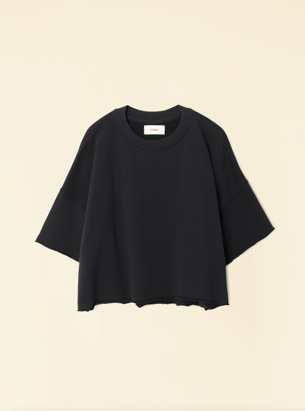 The Romeo Sweatshirt by Xirena, an oversized black garment with drop shoulders and wide sleeves, is displayed against a pale beige background. Made from ultra-soft fleece, this sweatshirt features a slightly rugged, unfinished hem at the bottom and a simple round neckline. A small white label is visible inside the collar.