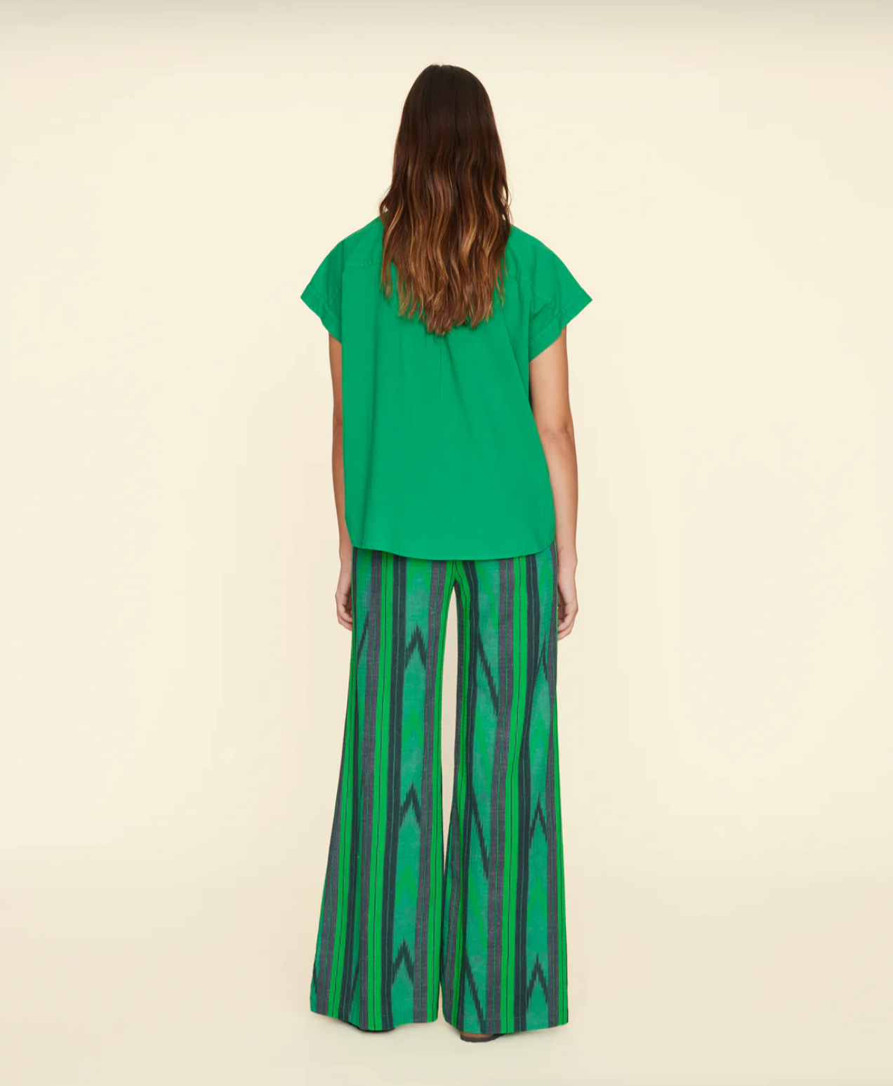 A person with long brown hair stands with their back to the camera, wearing a short-sleeved green top and the Xirena Pippen Pant, which features an ikat stripe pattern in green and blue and boasts an elasticized waist for comfort. The background is a plain light beige.