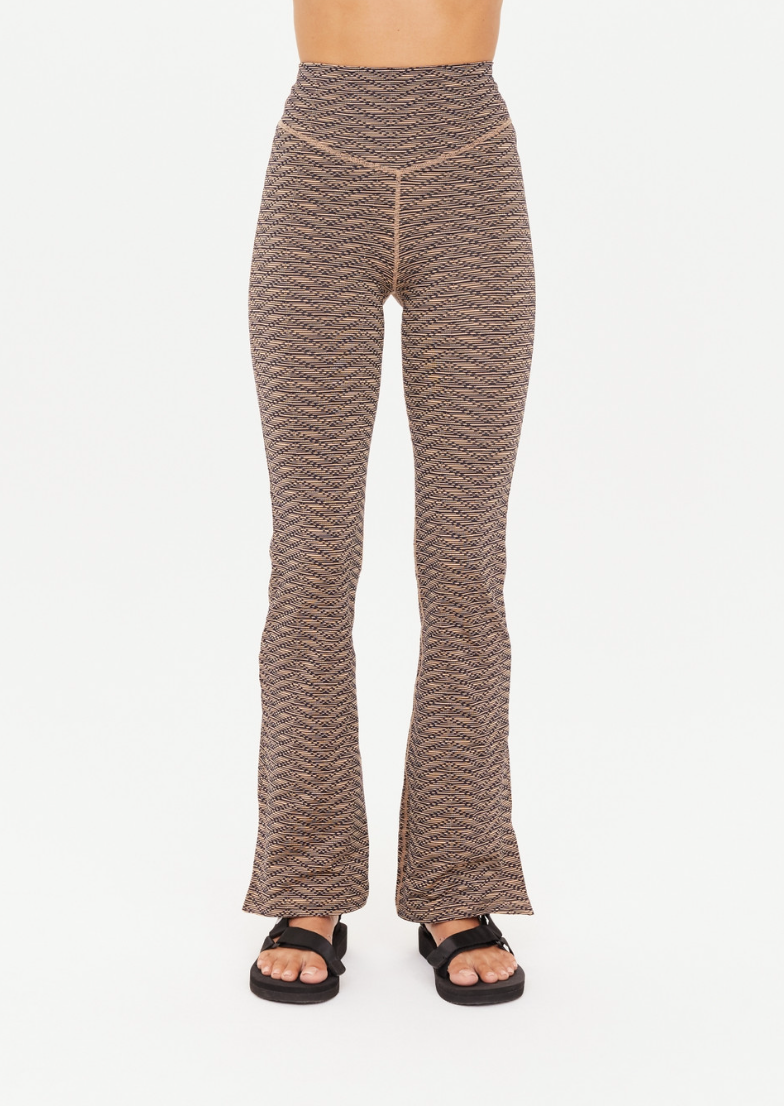 A person is wearing the LIBERTINE FLORENCE FLARE pants by The Upside, which are high-waisted with a textured wavy pattern in neutral tones. The pants feature a subtle gold zig-zag design and flare out slightly at the bottom. The individual is also wearing black open-toe sandals. The background is plain white.