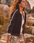 A woman with curly brown hair walks outdoors in a nature setting, wearing The Upside's CASTILLON KASIA JACKET, which is black with blue and white stripes on the sleeves. She pairs the jacket featuring welt pockets with a white top and metallic silver shorts. She accessorizes with gold earrings and bracelets. A logo reads "Sustainably Focused.