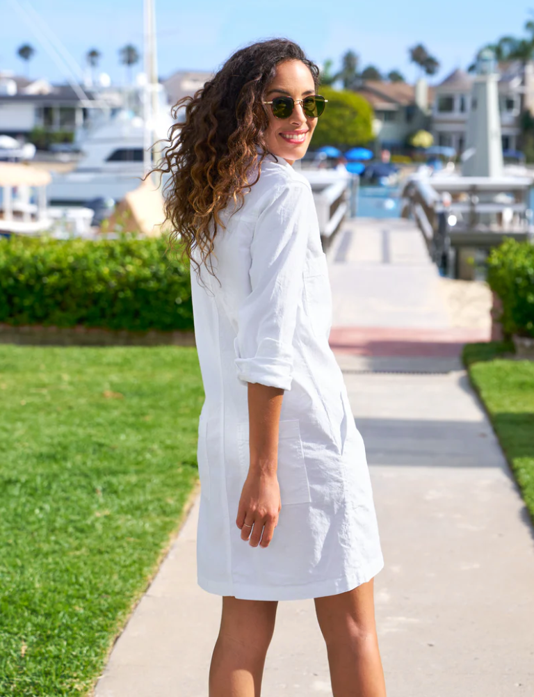 A woman with curly hair, wearing sunglasses and a white Southern Dress from Frank &amp; Eileen, is standing on a pathway by the water in Southern Ireland. She is looking back over her shoulder and smiling. The background includes boats, greenery, and houses.