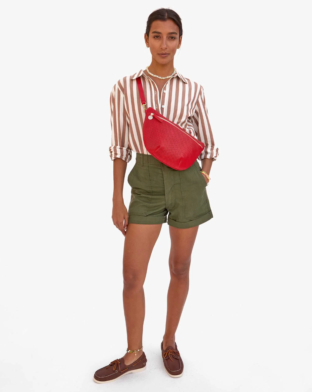 A person stands against a plain background, wearing a striped shirt, olive green shorts, and brown boat shoes. They have the Grande Fanny Perf from Clare Vivier with an adjustable strap slung over one shoulder and sport delicate jewelry including a necklace and bracelet for a stylish hands-free look.