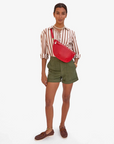 A person stands against a plain background, wearing a striped shirt, olive green shorts, and brown boat shoes. They have the Grande Fanny Perf from Clare Vivier with an adjustable strap slung over one shoulder and sport delicate jewelry including a necklace and bracelet for a stylish hands-free look.