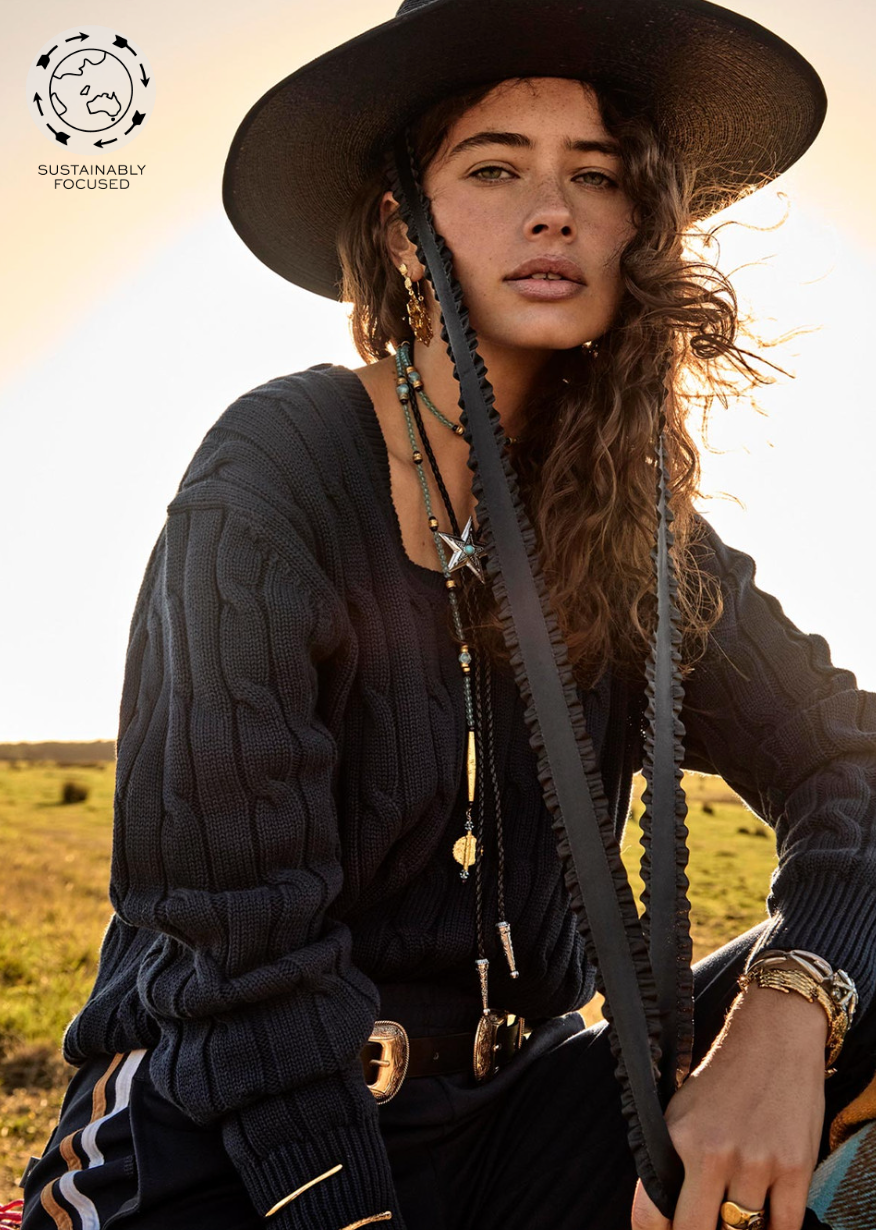 A woman with long, wavy hair wears a wide-brimmed black hat and a dark SEYMOUR KNIT SWEATER from The Upside, featuring a V-shaped neckline. She is adorned with various necklaces and gazes directly at the camera. The background showcases a grassy field bathed in sunset light, while the top left corner displays a circular logo with &quot;Sustainably Focused.