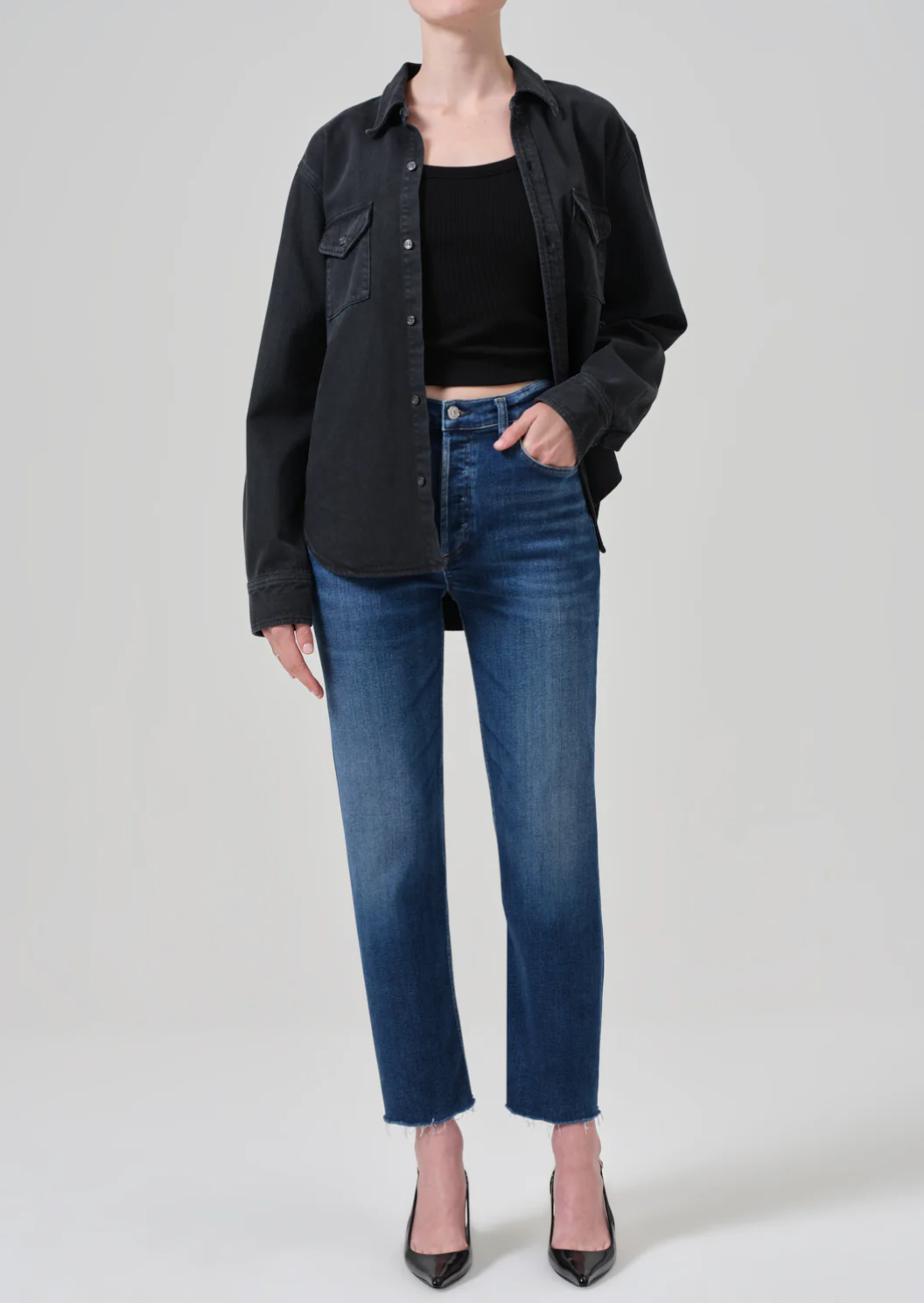 A person wearing a black button-up shirt, a black tank top, Citizens Of Humanity/AGOLDE palma straight jeans in ambry, and black high heels stands against a plain grey background. The shirt is worn open, and one hand is placed inside the front pocket of the jeans. The person's head is not visible.